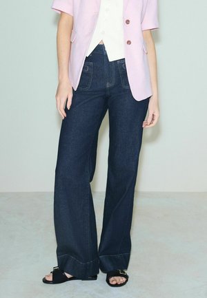 Inimene, kes kannab tumedaid laiade säärega teksapükse, heledat roosat jopet pruunide nööpidega, valget särki ja musti avatud varbaga sandaale, seisab tavalise tausta ees.