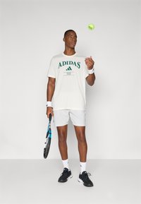 Camiseta branca com o logotipo "ADIDAS" verde, calções cinzentos, sapatilhas pretas, segurando uma raquete de ténis e a lançar uma bola de ténis verde brilhante.