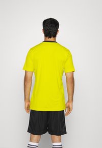 Camiseta atlética amarilla de mangas cortas, con un cuello negro, combinada con pantalones cortos de malla negros. Diseño simple y funcional.