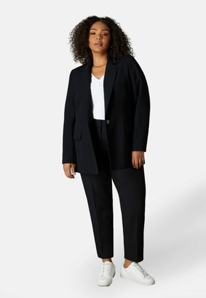 Blazer marine avec deux grandes poches avant, associé à un pantalon marine ajusté, un top blanc en V et des baskets blanches. Texture de tissu lisse.