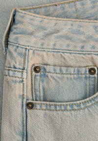 Gros plan sur un jean en denim bleu clair montrant la ceinture, la passants de ceinture et la poche avant avec des détails de rivets et des coutures jaunes.
