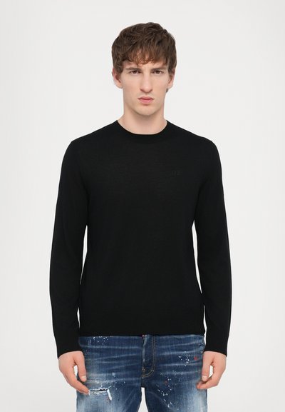 Pull en tricot noir à manches longues avec un col rond, présentant un logo subtil sur la poitrine, associé à un jean en denim déchiré.