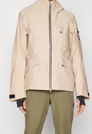 Veste à capuche beige avec fermeture éclair sur le devant, poches latérales inclinées et poignets réglables, associée à un pantalon vert olive.