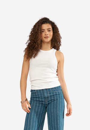 Femme aux cheveux bouclés portant un haut blanc sans manches et un jean bleu à fines rayures, debout devant un fond uni.
