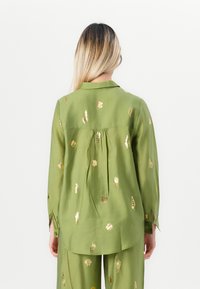 Frau mit langen blonden Haaren trägt ein grünes Oberteil und eine passende Hose, die mit goldenen Muschelmustern verziert ist, und steht vor einem weißen Hintergrund.