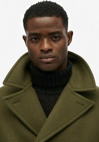 Abrigo de lana verde oliva con amplios reversos, superpuesto sobre un cuello de tortuga negro de canalé. Textura suave y silueta estructurada.
