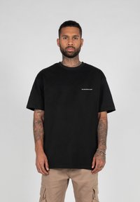 Schwarzes Baumwoll-T-Shirt mit kurzen Ärmeln, das einen Rundhalsausschnitt und ein kleines weißes Logo auf der linken Seite hat. Lockerer Schnitt und glatte Textur.