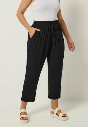 Mujer vistiendo pantalones negros con cordón y detalle de encaje en el lateral, top blanco sin mangas y sandalias blancas de plataforma, de pie sobre suelo beige.