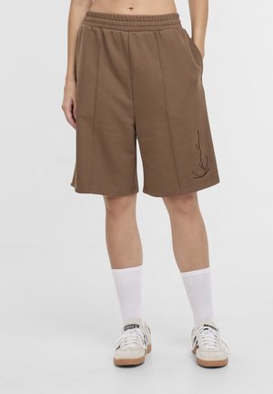 Személy barna térdig érő rövidnadrágot visel, rugalmas derékszíjjal és oldalzsebekkel, fehér zoknival és fehér Adidas tornacipőkkel.