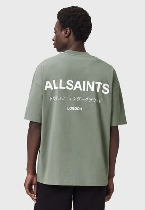 Homme de dos portant un t-shirt vert olive surdimensionné avec le texte blanc « ALLSAINTS », des caractères japonais et « LONDON » au dos.