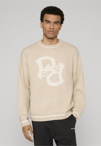 Beige gebreide trui met een groot wit grafisch logo aan de voorkant, geribde halslijn en manchetten, zachte textuur, relaxte pasvorm, gecombineerd met zwarte broek.