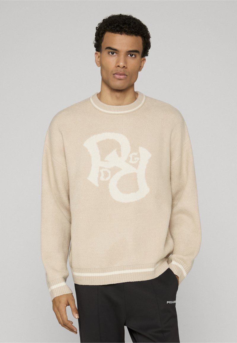 Beige gebreide trui met een groot wit grafisch logo aan de voorkant, geribde halslijn en manchetten, zachte textuur, relaxte pasvorm, gecombineerd met zwarte broek.