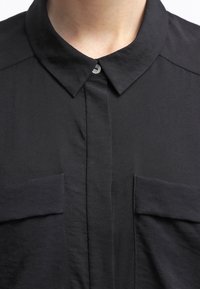 Czarna koszula zapinana na guziki z kołnierzykiem, wyposażona w dwie kieszenie na piersi i gładką fakturę. Prosty design bez wzorów czy akcentów.