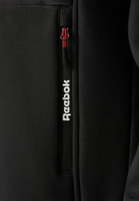 Czarna sportowa kurtka z kieszenią na zamek i widocznym białym logo "Reebok". Gładka tkanina z nowoczesnym designem. Czerwony akcent na zamku.