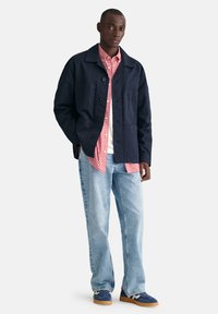 Marineblaue Baumwolljacke mit Knopfverschluss und zwei Vorderaschen, kombiniert mit einem roten Karoshirt und einem weißen T-Shirt, dazu hellblaue Jeans.