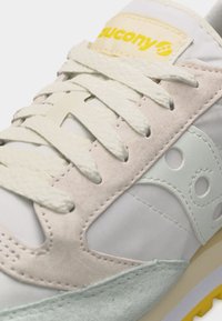 Κοντινή λήψη λευκού και μπεζ αθλητικού παπουτσιού Saucony με κρεμ κορδόνια, σουέτ επενδύσεις και κίτρινο λογότυπο στη γλώσσα.