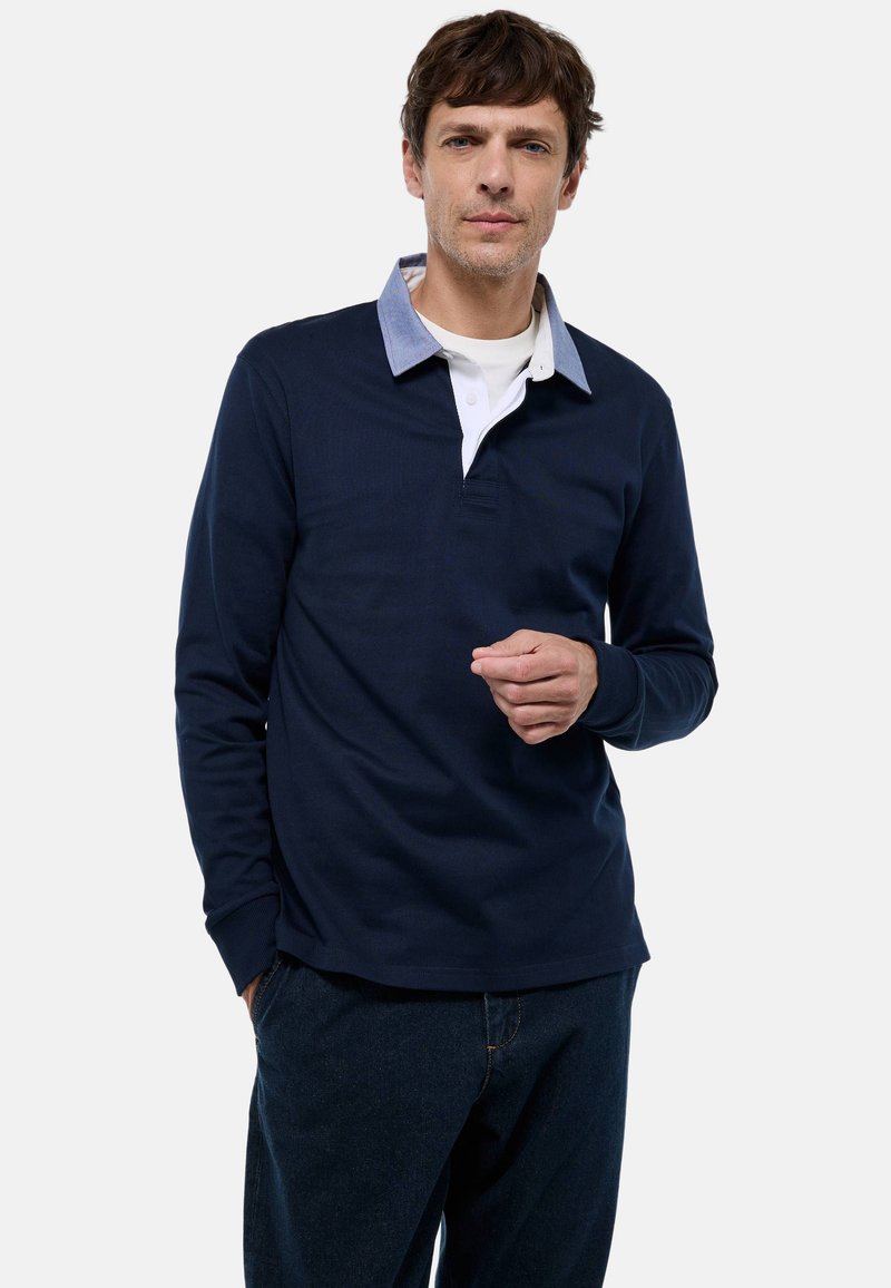 Polo shirt à manches longues marine avec col bleu clair, patte de boutonnage blanche et tissu texturé. Le modèle porte un jean bleu foncé. Design simple.