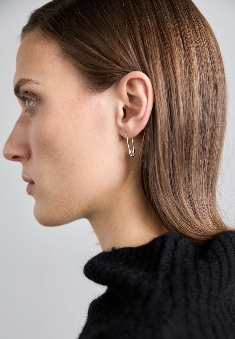 Sidabrinės auskarai-žiedai, forma primenantys dideles saugumo segtukus, pasižymi elegantišku dizainu su poliruotu paviršiumi. Matoma tekstūruota tamsi megztinį.