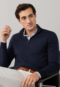 Muž sedící na židli, oblečený v námořnicky modrém svetru na zip, světle modré košili s límečkem, bílých kalhotách a hnědém koženém opasku, dívající se stranou.