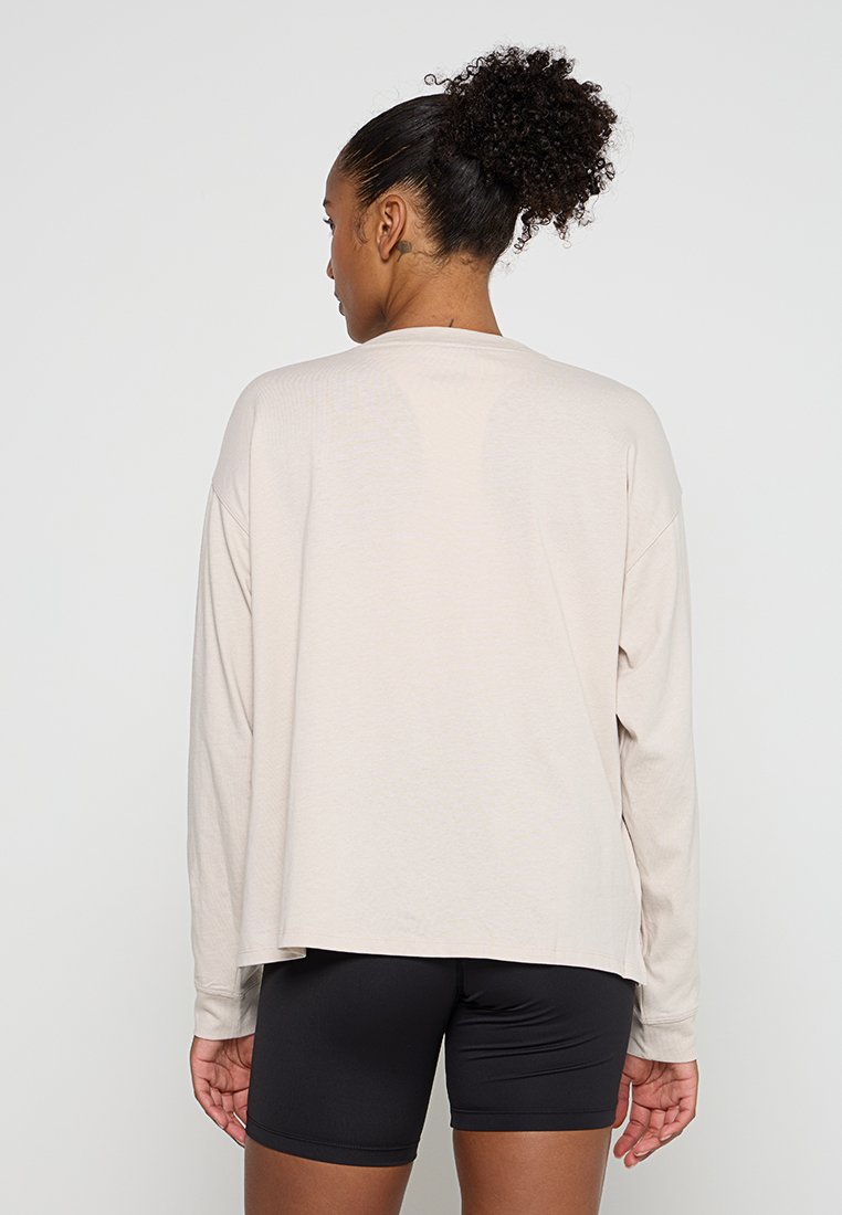 Frau mit lockigem Haar, das zu einem Dutt gebunden ist, trägt ein lockeres, langärmliges beiges Oberteil und enge schwarze Shorts. Sie steht mit dem Rücken zu einem schlichten Hintergrund.