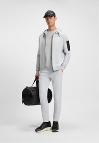 Férfi világosszürke melegítőben és fekete sapkában, fekete sporttáskával a kezében, egy sima fehér háttér előtt áll.