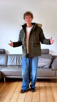 Sötétzöld puffdzseki, szőrmebéléssel ellátott kapucnival, gombos elülső résszel és két elülső zsebbel; világoskék, bőszárú farmernadrággal és fekete cipővel párosítva.