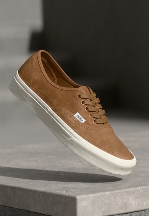 Brun ruskind Vans sneaker med hvid gummisål og snørebånd, placeret i luften over en grå betonoverflade.
