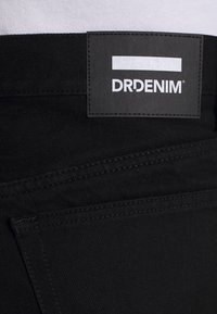 Jeans de mezclilla negros con una etiqueta visible de "DRDENIM", que cuentan con una tela texturizada, costuras limpias y detalles de bolsillos tradicionales.
