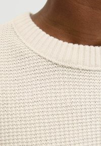 Light cream-knit sweater with ribbed collar. Textured weave pattern and soft fabric create a cozy appearance. Close-up view highlights details.