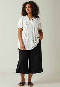 Weißes Oberteil mit Zebra-Streifen und gerafften Ärmeln, kombiniert mit schwarzen, weit geschnittenen Hosen und schwarzen Slidesandalen. Lässiges, leichtes Outfit.