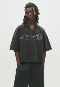Black oversized short-sleeve polo shirt with a v-neck, featuring a graphic print on the front and a relaxed fit. Smooth cotton fabric.
