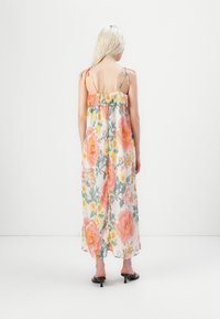 Femme aux cheveux blonds portant une robe maxi sans manches à fleurs orange et jaunes, debout devant un fond blanc uni.