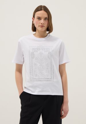 Femme aux cheveux bruns courts portant un T-shirt blanc orné d'un motif floral détaillé en clous argentés, associé à un pantalon noir.