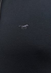 Ciemny materiał z małym wyszywanym emblematem biegnącego konia na środku, ukazujący szczegółową teksturę dzianiny.