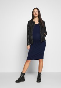 Casaco de couro preto sobre um vestido justo azul-marinho até aos joelhos. Usado com botas de tornozelo pretas chunky com fivelas. Texturas suaves.