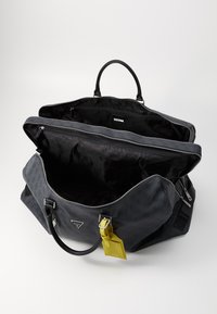 Sac de sport noir avec fermeture éclair, en matériau texturé, dotée de deux poignées et d'une étiquette de bagage jaune. Intérieur spacieux avec doublure logo.