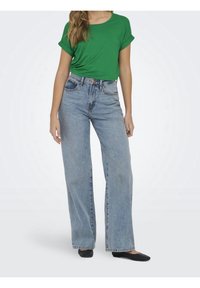 Femme portant un haut vert à manches courtes rentré dans un jean large taille haute bleu clair et des chaussures plates noires, debout sur fond blanc.
