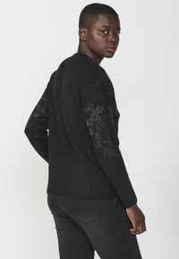 Maglione nero con finitura testurizzata, orlo a coste e dettagli floreali sulle maniche. Abbinato a jeans scuri, modello rivolto leggermente di lato.