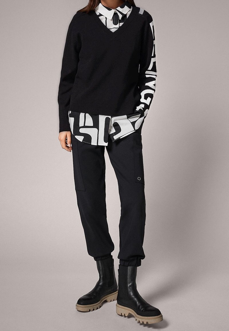 Personne portant un pull noir à col en V sur une chemise à motifs noir et blanc, un pantalon cargo noir et des bottes noires avec des semelles beiges.