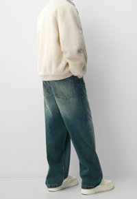 Chaqueta de forro polar en blanco roto, con una superficie texturizada. Vaqueros de denim azul holgados con un efecto desgastado, combinados con zapatillas crema.