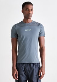 Pelēka īsām piedurknēm sporta krekls, izgatavots no gludas, elastīgas auduma, ar ribota plecu dizainu un baltu "CALVIN KLEIN" logotipu.