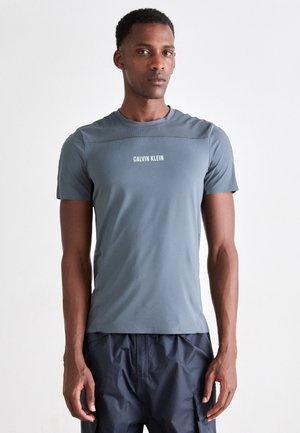 Grå kortermet treningsskjorte laget av et glatt, elastisk materiale, med et rillet skulderdesign og hvit "CALVIN KLEIN" logo.