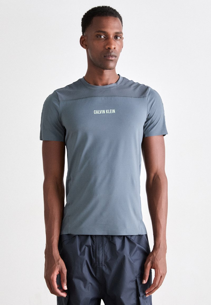 Pelēka īsām piedurknēm sporta krekls, izgatavots no gludas, elastīgas auduma, ar ribota plecu dizainu un baltu "CALVIN KLEIN" logotipu.