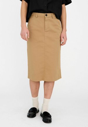 Person iført en beige knælængde nederdel, sort skjorte med knapper, hvide sokker og sorte loafers, stående mod en ensfarvet hvid baggrund.