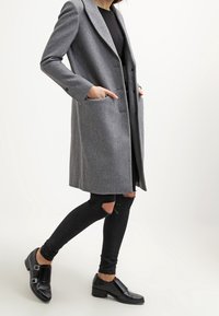 Cappotto lungo in misto lana grigio con revers, due tasche frontali e vestibilità rilassata. Abbinato a jeans skinny neri strappati e scarpe nere.