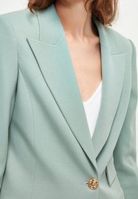 Světlý tyrkysový blazer vyrobený z texturované látky má hluboký výstřih do V, strukturovaná ramena a zlatý knoflík jako akcent vpředu.