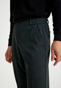 Dunkelgraue, taillierte Hose mit strukturiertem Finish, elastischem Bund, seitlichen Taschen und einem einzelnen Knopfdetail am Bund.