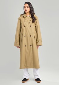 Tan waterproof trench coat with a double-breasted design, large buttons, side pockets, and an adjustable drawstring hood.