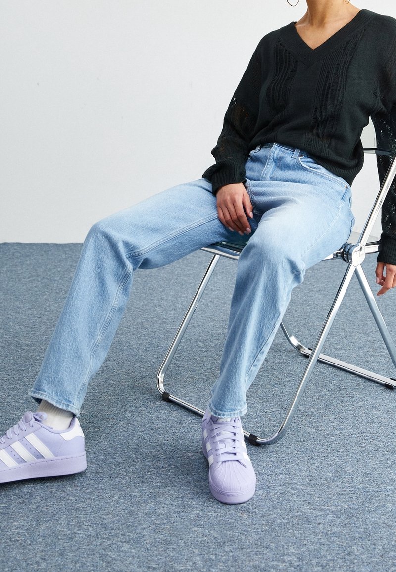 Černý pletený svetr s výstřihem do V, světle modré džíny s vysokým pasem a levandulové tenisky s bílými pruhy. Sedí na chromov é židli.