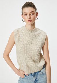 Beige mouwloze gebreide trui met een gestructureerd kabelpatroon, geribbelde ronde halslijn en een ontspannen pasvorm, gecombineerd met lichte denim spijkerbroek.
